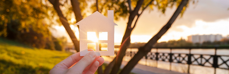 Banner with hand that holds a paper house against the background of the sunset sky. The sun is setting over the horizon. The concept of your own home, personal space and mortgage. High quality photo