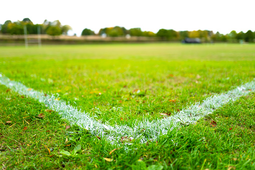 Shallow focus of the corner of painted white lines seen on a public school's rugby field. A distant rugby goal can be seen.