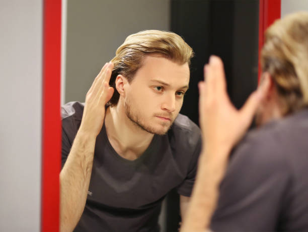 istock young man looking in the mirror,combing his hair,looking at problems on face 1433613455