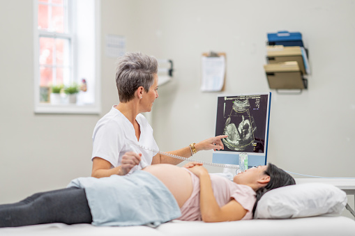 A female healthcare professional sits beside a pregnant patient as she lays out on the exam table for an ultrasound.  The technician is showing the patient her baby on the screen as she conducts the appointment.