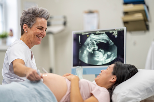A female ultrasound technician holds the wand to her pregnant patients belly as she conducts the appointment.  She is wearing white scrubs and is smiling at the patient as she checks on the baby.