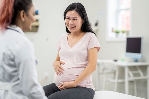 A young pregnant woman, of Asian decent, sits up on an exam table as she talks with her doctor during a prenatal check-up.  She is dressed casually and holding her belly as the two talk.