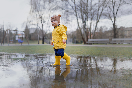 Happy little boy in yellow raincoat jumping in puddle after rain in cold autumn day.