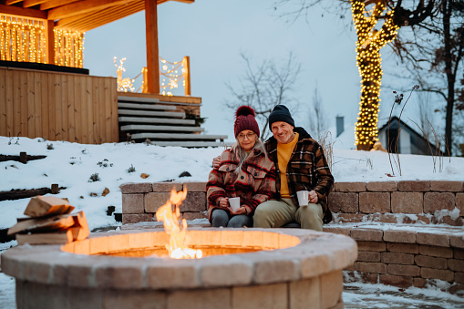 Senior couple sitting and heating together at outdoor fireplace during winter evening.