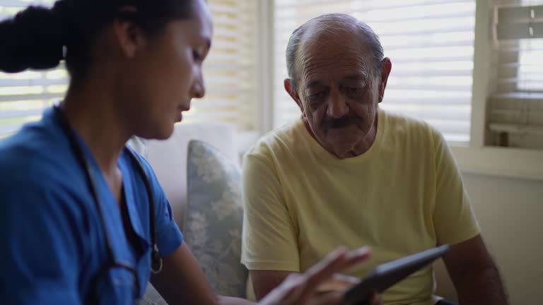 Mid adult nurse using digital tablet while talking to a senior man in a consultation in the living room of a nursing home