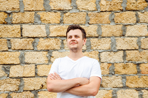 Young caucasian 34 years old man in white t-shirt with cross arms chest on Stone bricks from limestone laid background. Waist up lifestyle Looking away unshaven man portrait. Entrepreneur, businessman