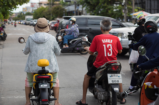 Thai people are standing on motorcyles at stoplight and junction. A senior woman with hat is sitting on electric motorcycle. All others are using gasoline engine motorcycles. On junction traffic is ruling