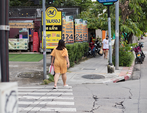 Young thai woman in yellow colored casual clothing is crossing crosswalk and sidewalk of street Wanghin Road in Bangkok ladprao. In background is a banner. In far right backgrond are other pedestrians