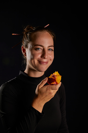 young russian girl looking at camera smiling with nectarine fruit at hand wearing black dress and black background