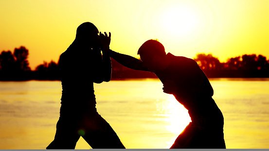 Two dark male figures, at sunrise, against the light, boxing, fighting in sparring, training in a pair of techniques of strikes. On sandy beach, in cargo port, near water, in summer. High quality photo