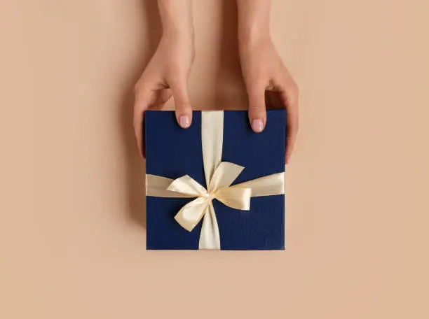 Female hands with natural manicure holding blue gift box with light golden ribbon on trendy beige background. Xmas and New Year postcard design. Black Friday sales, Birthday celebration party concept