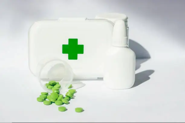 A small first aid kit with pills and green cross on the lid. There are jars of medicines nearby. All this on a white background
