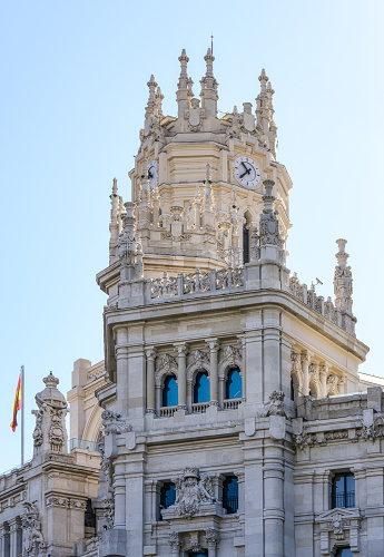 Madrid, Spain - September 25, 2022: Low-angle view of the upper part of a palace. The building has a clock tower at its upper section, and the Spanish flag is on one side of the building.