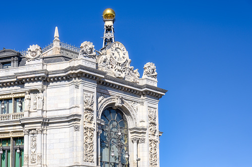 Madrid, Spain - September 25, 2022: Low-angle view of the upper part of a bank. Beautiful sculptures are on the building, and a tower-like structure holding a gold ball sits atop the building.