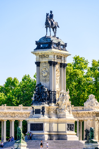 Madrid, Spain - July 18, 2022: People standing and sitting beside the monument to Alfonso VII. The structure comprises a splendid collonade with many sculptures encircling an equestrian sculpture of the king.