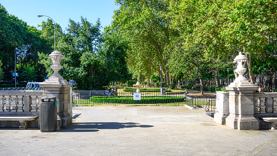 Madrid, Spain - July 18, 2022: People sitting on a bench inside a fenced public park.  There are several tall trees and manicured shrubs inside the park.