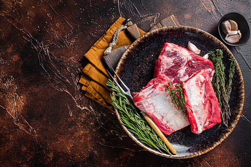 Uncooked Raw veal Short Ribs on rustic plate with rosemary. Dark background. Top view. Copy space.
