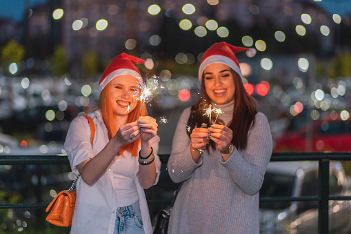 Waist-up portrait of two beautiful women wearing Santa hats and holding sparklers. They are celebrating Christmas/New Year.