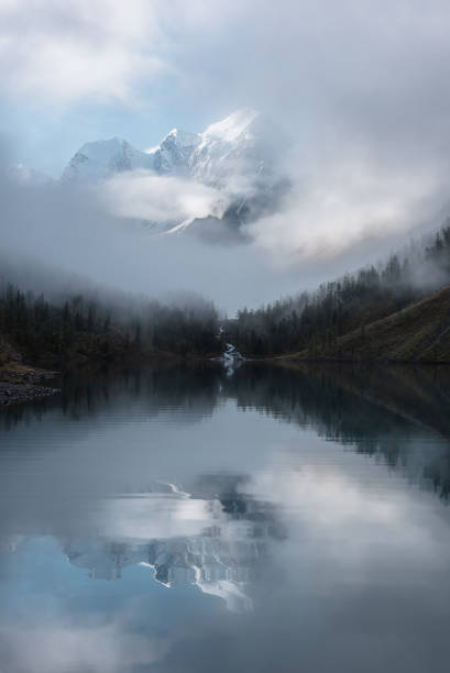 istock Tranquil scenery with snow castle in clouds. Mountain creek flows from forest hills into glacial lake. Snowy mountains in fog clearance, small river and coniferous trees reflected in calm alpine lake. 1433695632