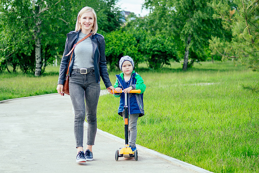 mother teaches the child young boy to ride on a scooter in a park on a background of green grass and trees