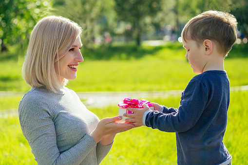 cute baby (son) gives a gift box to his beautiful blonde mother in the park on a background of green grass