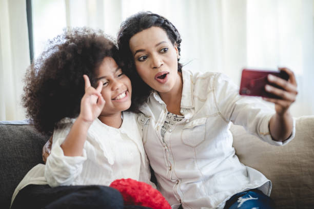 istock Cheerful young mother taking a selfie using smartphone with sister with curly hair while making funny facial and hand gestures sitting on couch at home 1433758997