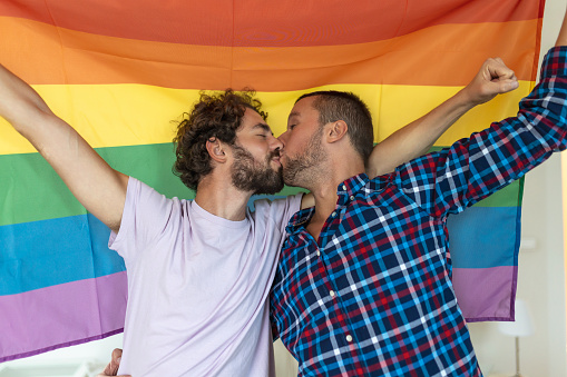 Two young gay lovers kissing each other affectionately. Two young male lovers standing together against a pride flag. Affectionate young gay couple sharing a romantic moment together.