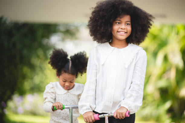 istock Two playful African sisters with curly hair enjoying and playing while riding scooter on empty lane surrounded by trees during day 1433769544