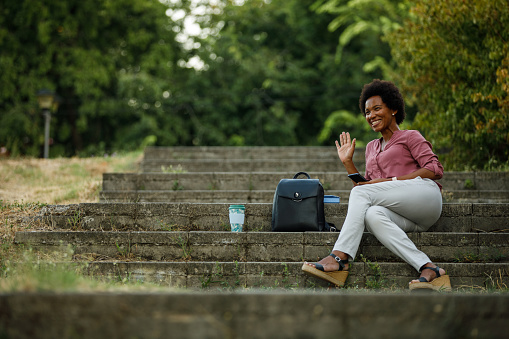 Woman waiving to a friend while resting outdoors after work, sitting on the stairs in the park.