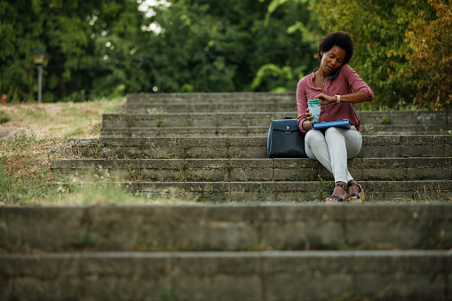 Businesswoman talking on the phone while sitting on the stairs outdoors in the park, checking time on her smart watch, having a coffee break.