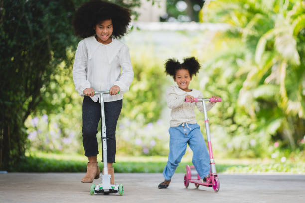 Two playful African sisters with curly hair enjoying and playing while riding scooter on empty lane surrounded by trees during day stock photo