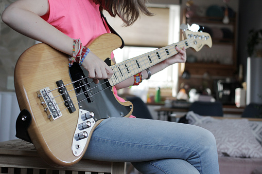 A young white girl with straight brown hair plays a light bass guitar sitting on a dresser at home guitar close-up