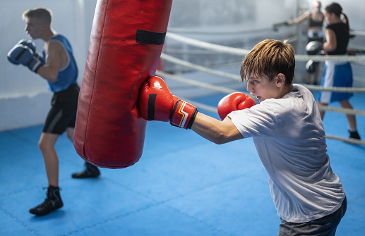 Teenage boy exercising at boxing with red boxing gloves on punching bag with another kids from boxclub. Combat sport and healthy lifestyle concept.