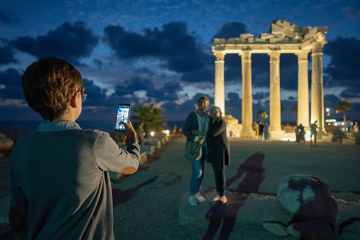 Young boy taking a photo of his grandmother and mother with his phone in front of the temple of Apollo in the ancient city side.