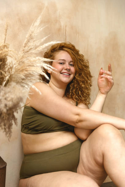 Beautiful woman in underwear Body positive model with redhead. Portrait of an attractive and voluptuous young woman posing in her lingerie against a beige background in studio plus size boho clothing stock pictures, royalty-free photos & images