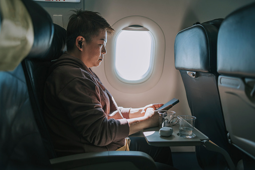 Asian Chinese woman using smart phone sitting on passenger window seat in airplane