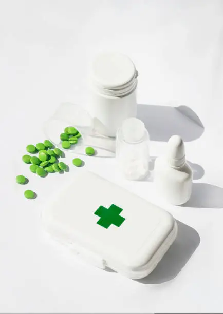 A small first aid kit with pills and green cross on the lid. There are jars of medicines nearby. All this on a white background
