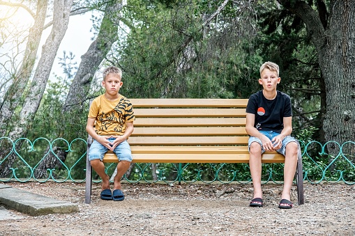 Siblings sitting on wooden bench look with upset and disappointed expressions. Brothers sit on different sides of bench in silence after argument