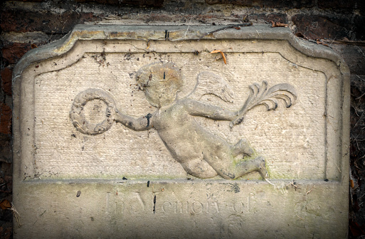 Part of an old gravestone in the churchyard of St Nicholas’ Chapel in King’s Lynn, Norfolk, Eastern England, showing a cherub carrying a wreath and a plume.