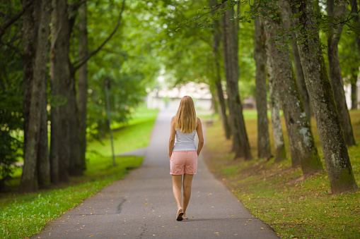 Young adult blonde woman slowly walking through alley of green trees on sidewalk at city park in warm summer day. Spending time alone. Peaceful atmosphere. Back view.