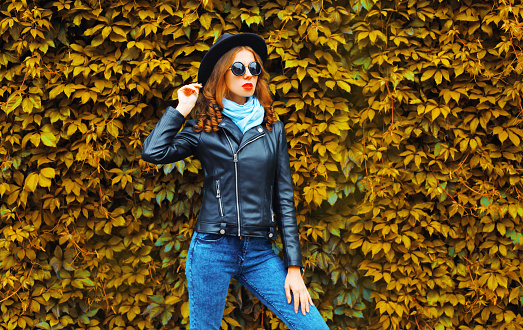 Portrait of beautiful young woman model wearing black round hat, leather jacket, sunglasses on leaves wall as background
