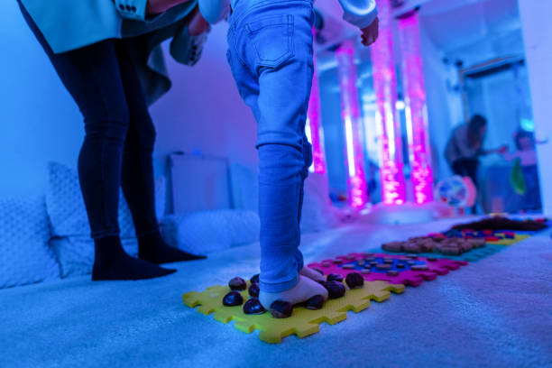 Therapist helping a young child living with cerebral palsy successfully improve her walking ability during therapy session in a sensory room, snoezelen. Texture walk, sensory exploration, motor skills. stock photo