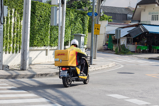 German parcel DHL driver on motorcycle is driving on street in Bangkok. Man is passing crosswalk and a junction and is turning left. Scene is in Bangkok Chatuchak