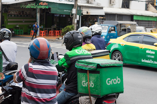 Thai male delivery person is standing with motorcyle in traffic and in street . Man is driving for company GRAB. Next to him another erson is on  motorcycle. On street is some traffic. Scene is on Wanghin Road in Bangkok Ladprao