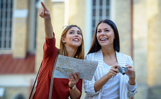 Two beautiful young women using the city map while exploring the locations and making a photos with camera. Concept of tourism