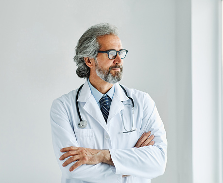 Waist-up portrait of physician with stethoscope around neck leaning against wall in inpatient facility hallway