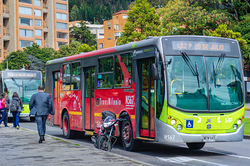 Bogota, Colombia - July 24, 2018: Two TransMilenio Public transportation at a bus stop on Calle 134 in the Latin American Andes capital city of Bogotá. Obviously one of them is running faster than he was supposed to! A motorcycle is illegally parked just ahead of the bus top. In the background are residential buildings and in the far background are the Andes mountains. The altitude at street level is 8,660 feet above mean sea level.  Horizontal Format.