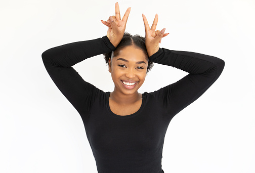 Portrait of funny young woman making bunny ears gesture. African American lady wearing black longsleeve having fun against white background. Humor concept