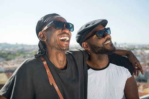 Close-up of happy African gay men spending time together. Two young bearded men in black glasses standing close on roof top hugging each other looking up. LGBT peoples love and relations concept