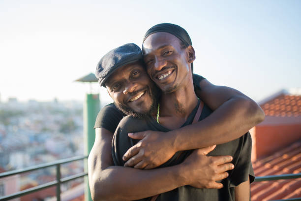 Close-up of happy African gay boyfriends looking at camera Close-up of happy African gay boyfriends looking at camera. Two bearded men in casual clothes standing together on roof top hugging taking photo on phone camera. LGBT peoples love, happiness concept two men same clothes stock pictures, royalty-free photos & images